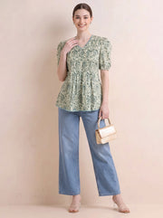 A woman modeling a light green and beige paisley print peplum kurti with puff sleeves, paired with light blue wide-leg jeans, a small beige handbag, and clear high-heeled sandals, set against a solid beige studio backdrop.
