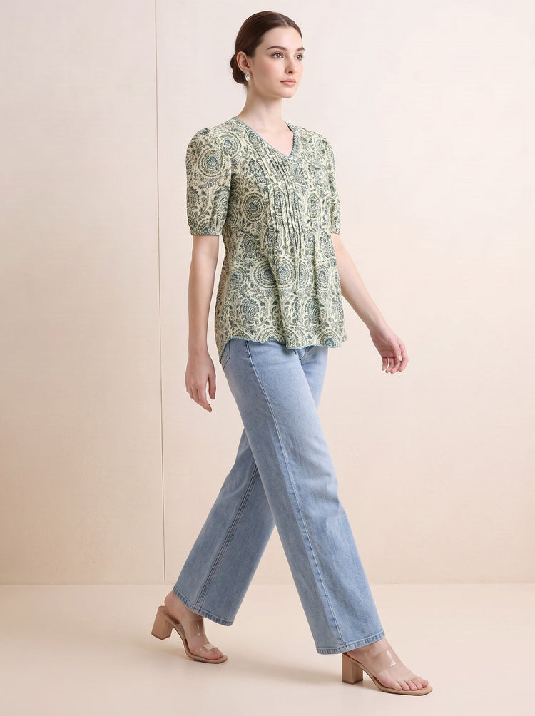 A woman modeling a light green and white patterned short-sleeve kurti with puff sleeves, paired with light blue jeans and beige block-heel sandals, set against a solid beige studio background.