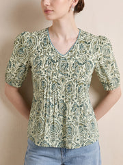 A woman wearing a short-sleeve V-neck kurti with a green and beige paisley pattern, paired with light blue jeans, standing against a neutral beige wall.