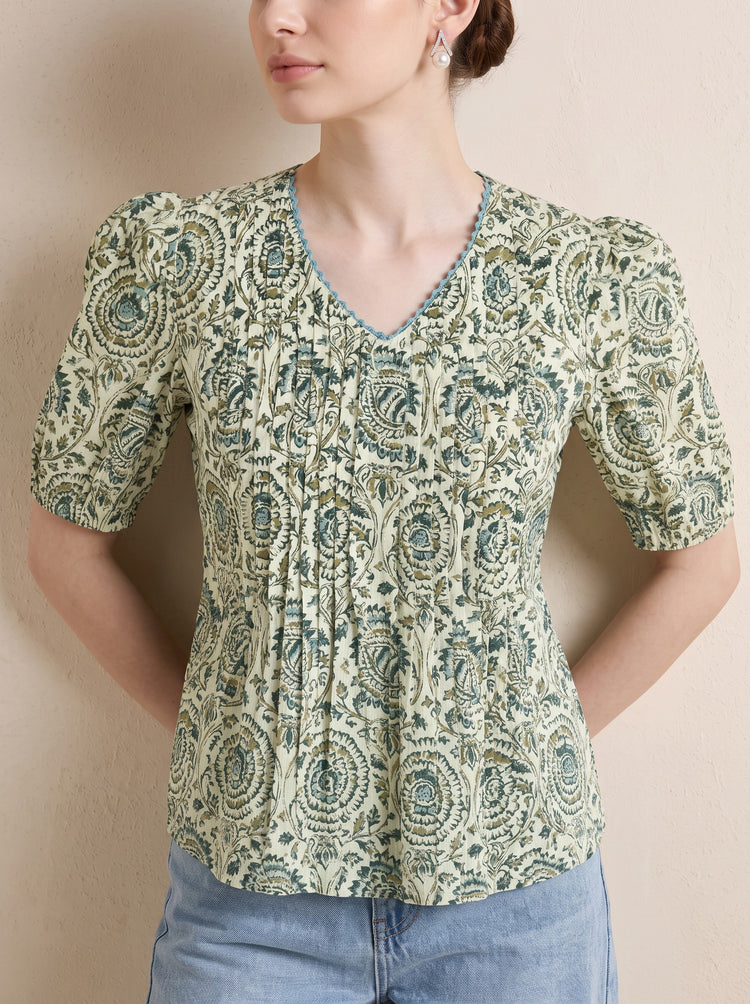 A woman wearing a short-sleeve V-neck kurti with a green and beige paisley pattern, paired with light blue jeans, standing against a neutral beige wall.