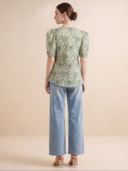 A woman is shown from the back wearing a short-sleeved top with a green and beige circular pattern, paired with light blue wide-leg jeans and clear high-heeled sandals, set against a solid beige studio background.