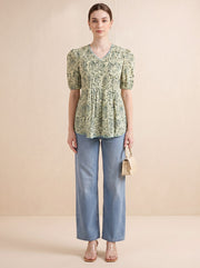 A woman stands against a plain beige studio background wearing a light green and cream patterned short-sleeve kurti with puff sleeves, paired with light blue wide-leg jeans and clear high-heel sandals. She holds a small beige handbag.