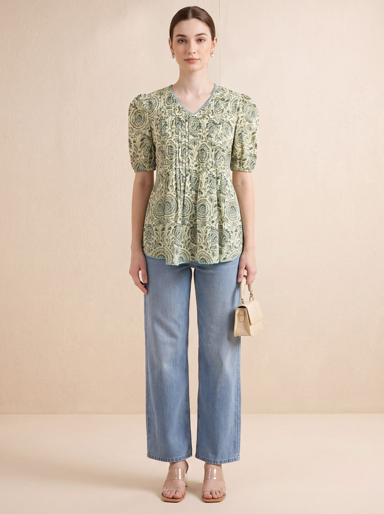 A woman stands against a plain beige studio background wearing a light green and cream patterned short-sleeve kurti with puff sleeves, paired with light blue wide-leg jeans and clear high-heel sandals. She holds a small beige handbag.