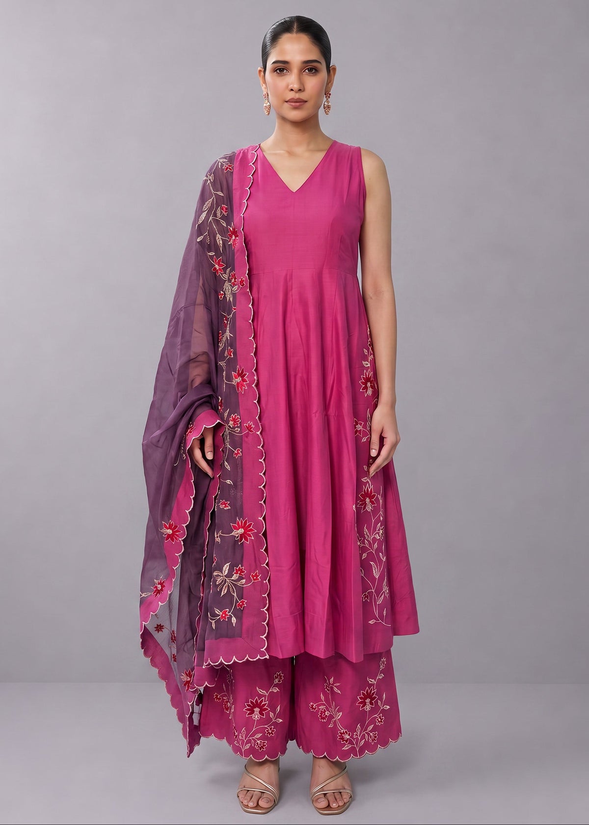 A woman modeling a magenta pink traditional Indian outfit consisting of a sleeveless V-neck kurta, matching palazzo pants, and a sheer purple dupatta with floral embroidery and scalloped borders, set against a solid gray studio background.