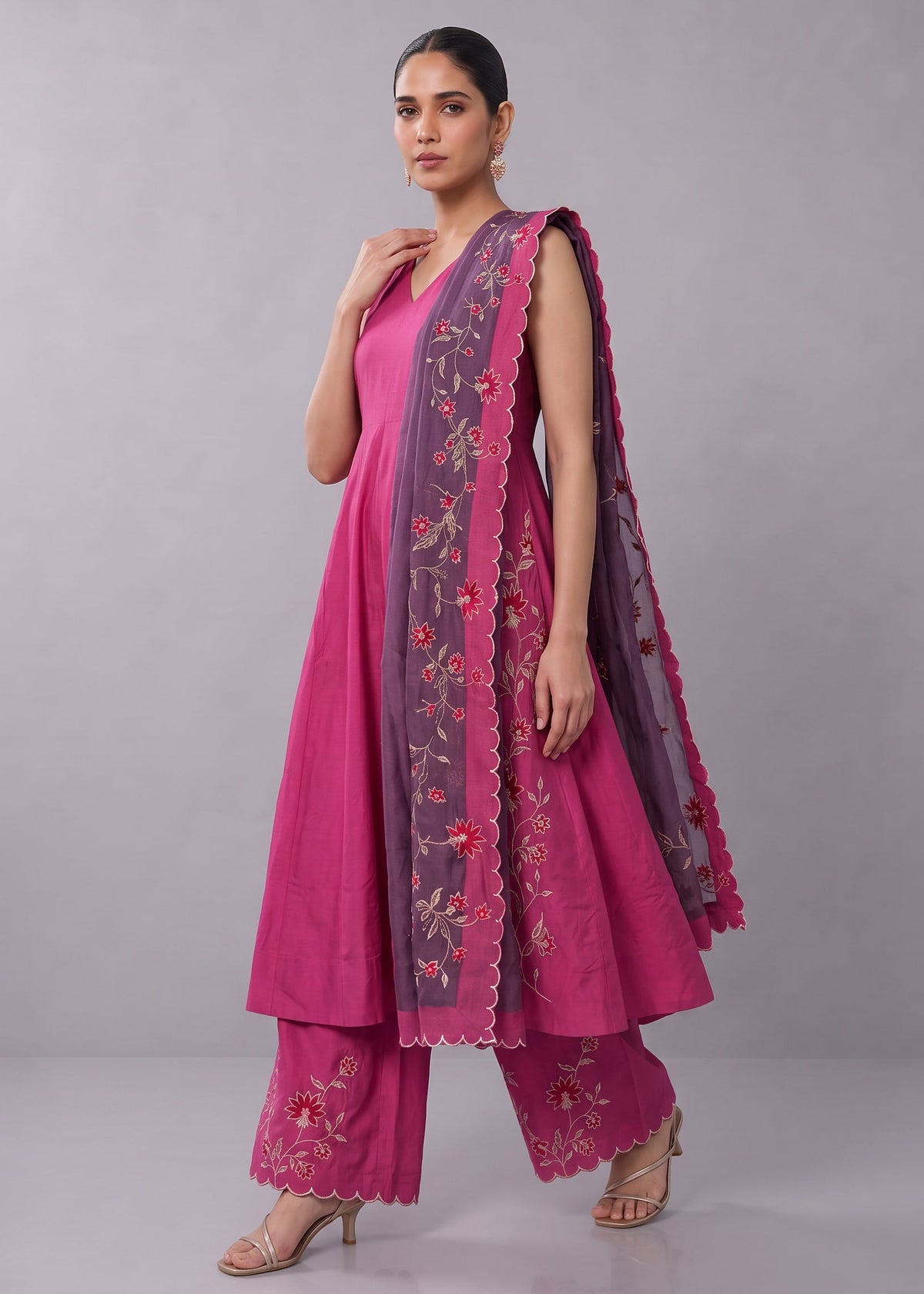 A woman models a traditional Indian salwar kameez suit in vibrant pink, featuring a pink kurta with a purple embroidered border, matching pink pants, and a sheer purple dupatta.