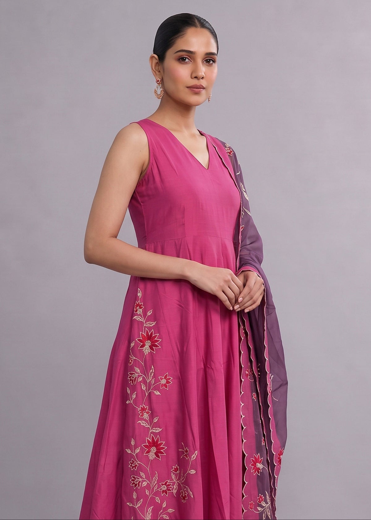 A woman modeling a sleeveless magenta pink Anarkali gown with floral embroidery and a matching purple dupatta against a solid gray studio background.