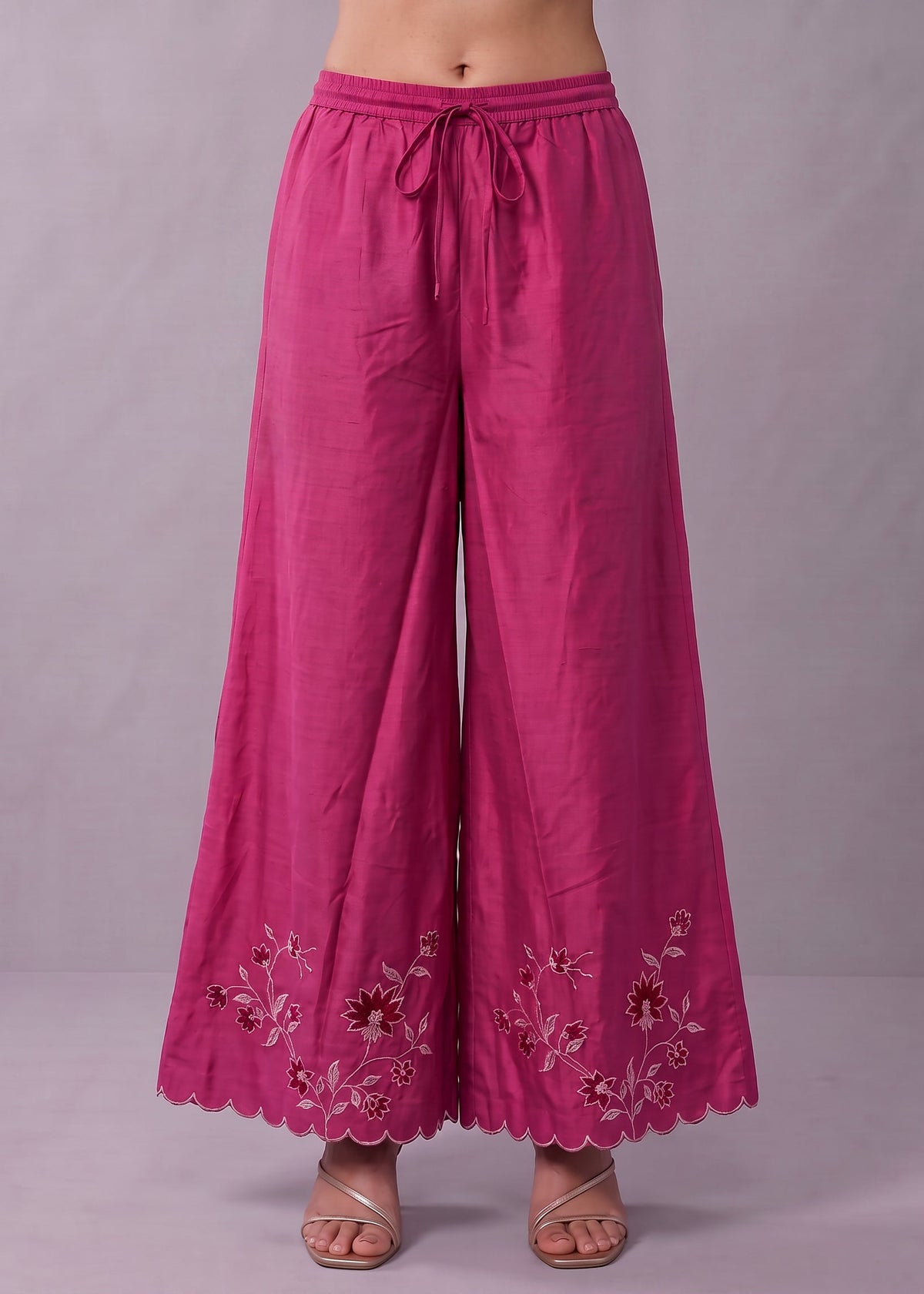A person wearing bright pink wide-leg pants with a drawstring waist and floral embroidery near the scalloped hem, shown against a plain light gray studio background.