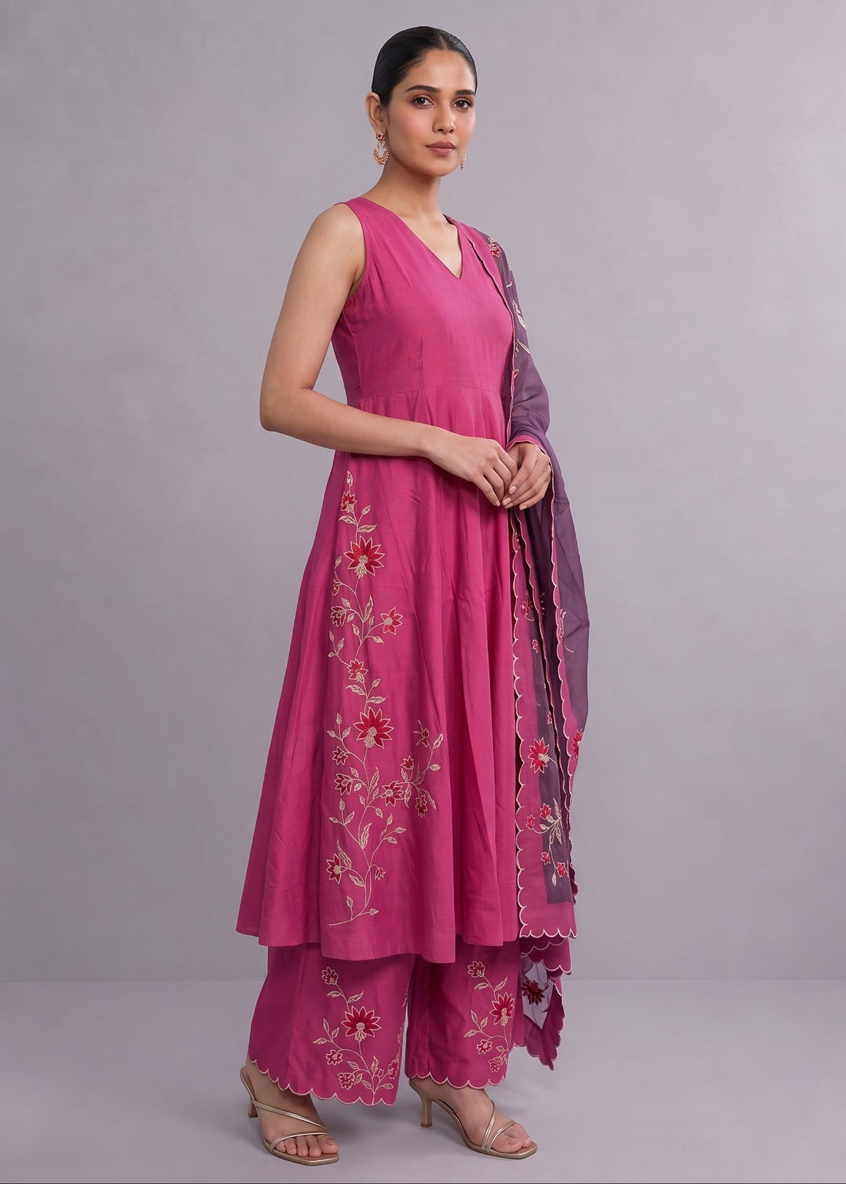 A woman modeling a magenta pink traditional Indian salwar kameez set with floral embroidery and a matching purple dupatta, standing against a solid gray studio background.