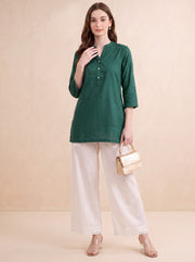 A woman modeling a dark green three-quarter sleeve kurta with a buttoned neckline, paired with wide-leg white trousers and holding a small metallic gold handbag, set against a warm beige studio backdrop.