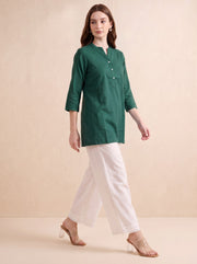 A woman modeling a dark green three-quarter sleeve kurta with a buttoned neckline, paired with white wide-leg trousers and clear high-heeled sandals, set against a warm beige studio backdrop.