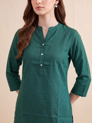 A woman wearing a dark green linen tunic with a split neckline and three-button placket, shown from the chest down against a neutral beige background.