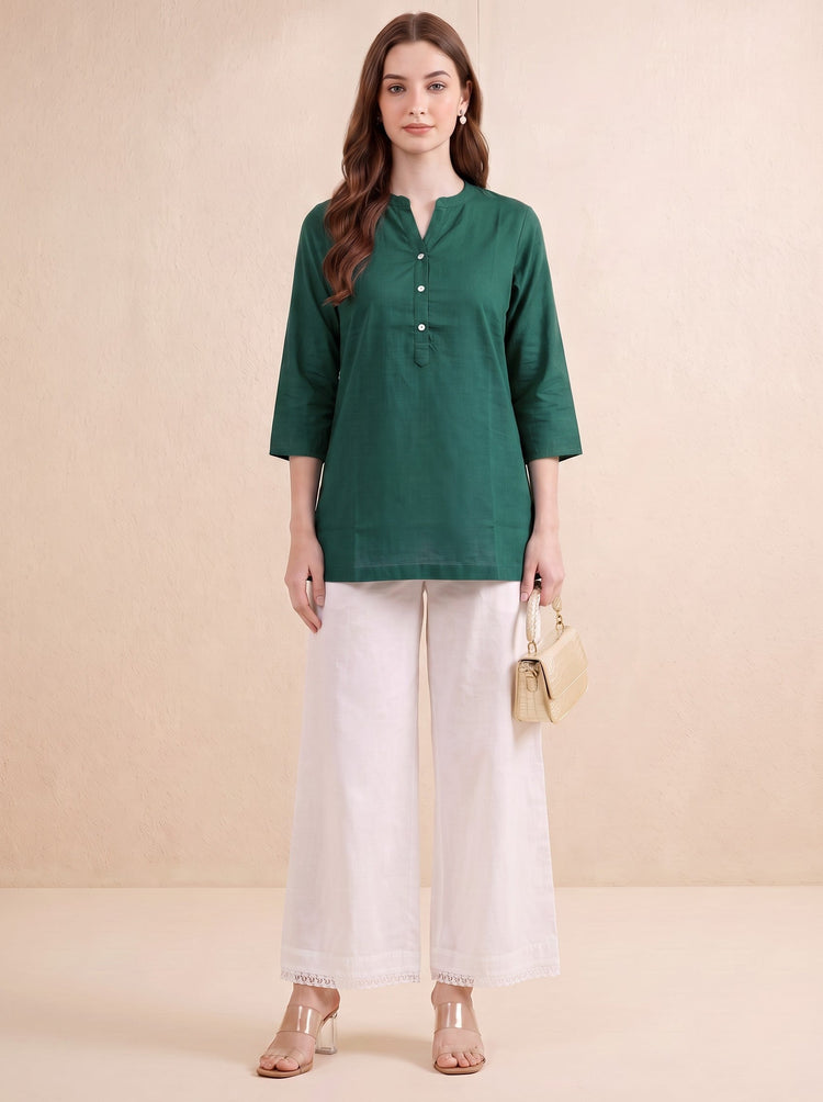 A woman stands against a warm beige studio backdrop wearing a dark green three-quarter sleeve kurta with a button placket, paired with wide-leg white pants and clear high-heeled sandals. She holds a small beige handbag.