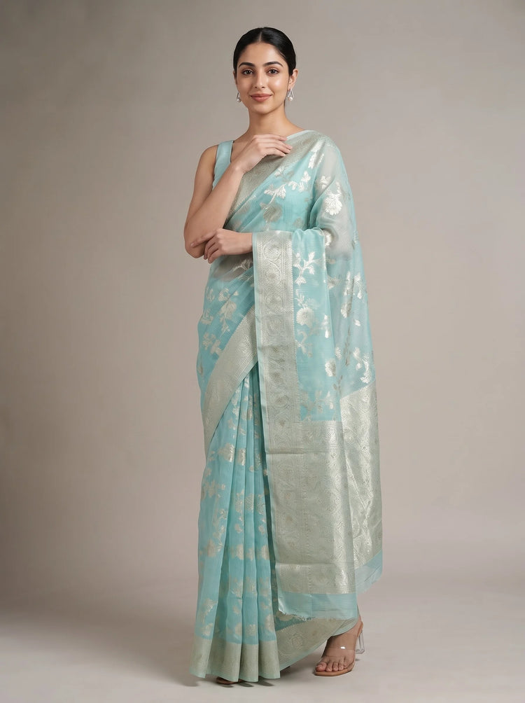 A woman modeling a light blue-green saree with metallic silver floral motifs and a wide silver border, photographed against a plain beige studio background.