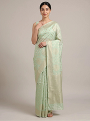 A woman modeling a light sage green saree with subtle woven motifs and a matching border, standing against a plain beige studio background.