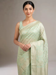 A woman modeling a light sage green silk saree with subtle gold zari motifs and a matching sleeveless blouse, set against a neutral gray studio background.