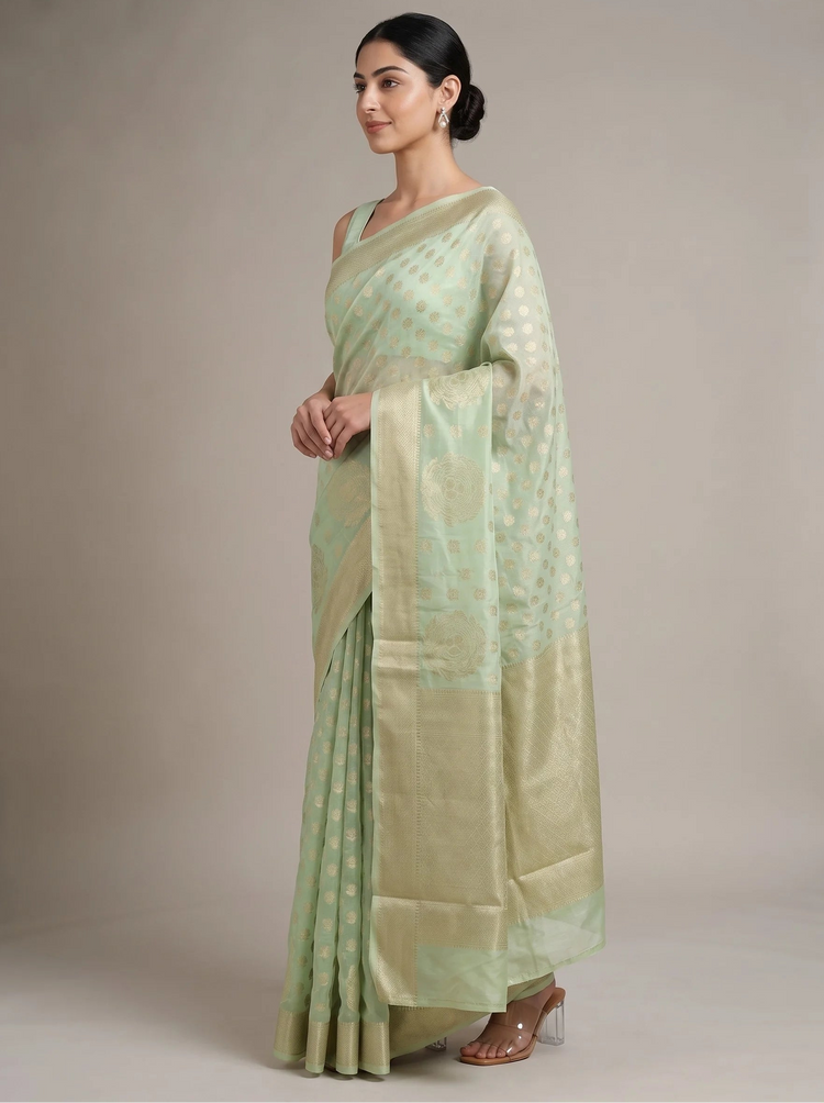 A woman modeling a light sage green saree with a subtle woven pattern and a wide border, standing against a neutral beige studio background.