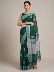 A woman modeling a dark green saree with a silver and light green border, featuring small metallic motifs and tassels, set against a plain beige studio background.