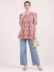 A woman models a short-sleeve V-neck top with a pink and beige circular floral pattern, paired with light blue wide-leg jeans, a small beige handbag, and clear high-heeled sandals, against a plain light beige studio background.