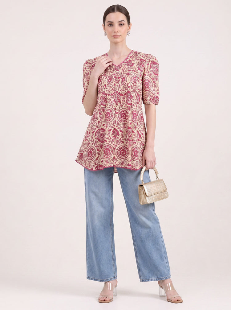 A woman models a short-sleeve V-neck top with a pink and beige circular floral pattern, paired with light blue wide-leg jeans, a small beige handbag, and clear high-heeled sandals, against a plain light beige studio background.