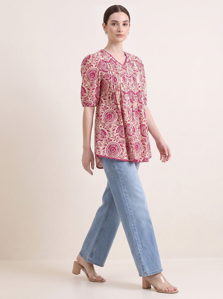 A woman modeling a short-sleeve V-neck kurti with a pink and beige floral paisley pattern, paired with light blue jeans and beige heeled sandals, against a solid light beige studio background.