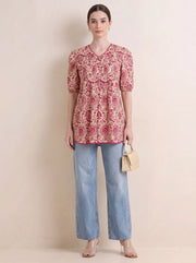 A woman stands against a plain beige studio wall wearing a short-sleeve V-neck top with a pink and beige paisley pattern, light blue wide-leg jeans, and clear high-heel sandals, while holding a small beige handbag.