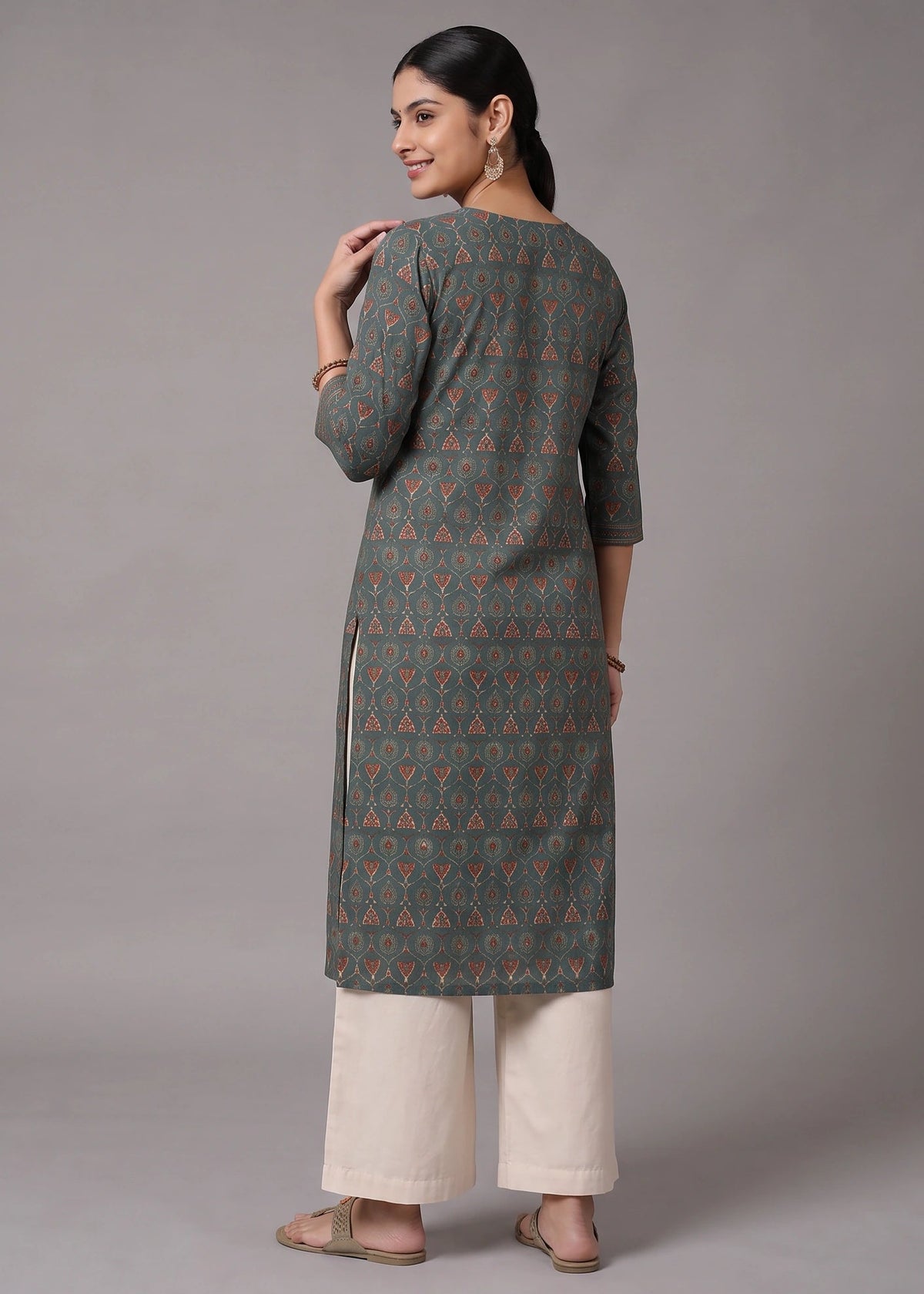 A woman modeling a long-sleeved kurta with a dark gray and orange geometric triangle pattern, paired with off-white wide-leg pants, shown from a rear three-quarter view against a neutral gray studio background.