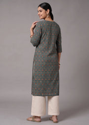 A woman modeling a long-sleeved kurta with a dark gray and orange geometric triangle pattern, paired with off-white wide-leg pants, shown from a rear three-quarter view against a neutral gray studio background.