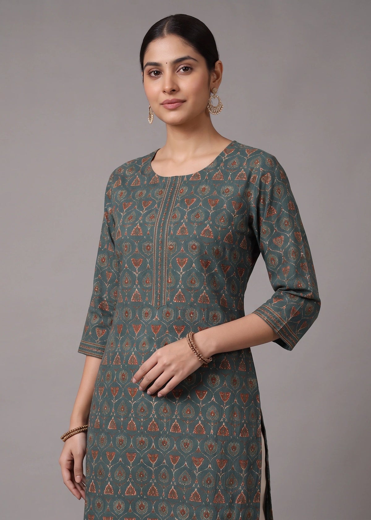 A woman modeling a three-quarter sleeve kurta in a muted teal-green color with a repeating orange and beige geometric block print pattern, set against a plain gray studio background.