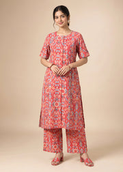 A woman modeling a red and blue patterned kurta set with matching pants and flat shoes, standing against a solid beige studio background.