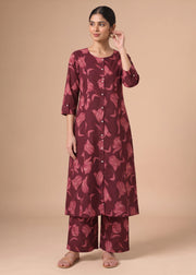 A woman modeling a three-piece kurta set in a deep maroon color with a pinkish floral pattern, consisting of a long button-up tunic, three-quarter sleeves, and matching wide-leg pants, set against a solid beige studio background.