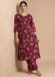 A woman modeling a three-piece Indian ethnic wear set consisting of a long-sleeve button-up kurta, pants, and flat sandals. The set is maroon with a pink floral pattern, set against a warm beige textured background.