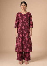 A woman modeling a three-piece kurta set in a deep burgundy color with a lighter pinkish-red floral pattern, standing against a solid beige studio background.