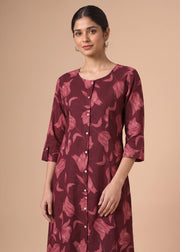 A woman modeling a long-sleeved button-front kurta in deep burgundy with a pinkish floral pattern, set against a solid beige studio background.