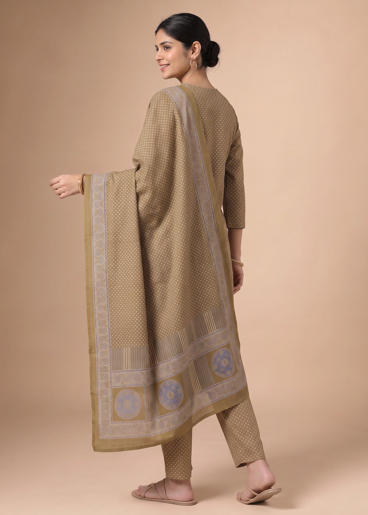 A woman modeling a traditional three-piece salwar kameez suit in beige with a subtle checkered pattern and a matching dupatta featuring blue and gold border designs, set against a solid beige studio background.