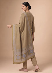 A woman modeling a traditional three-piece salwar kameez suit in beige with a subtle checkered pattern and a matching dupatta featuring blue and gold border designs, set against a solid beige studio background.