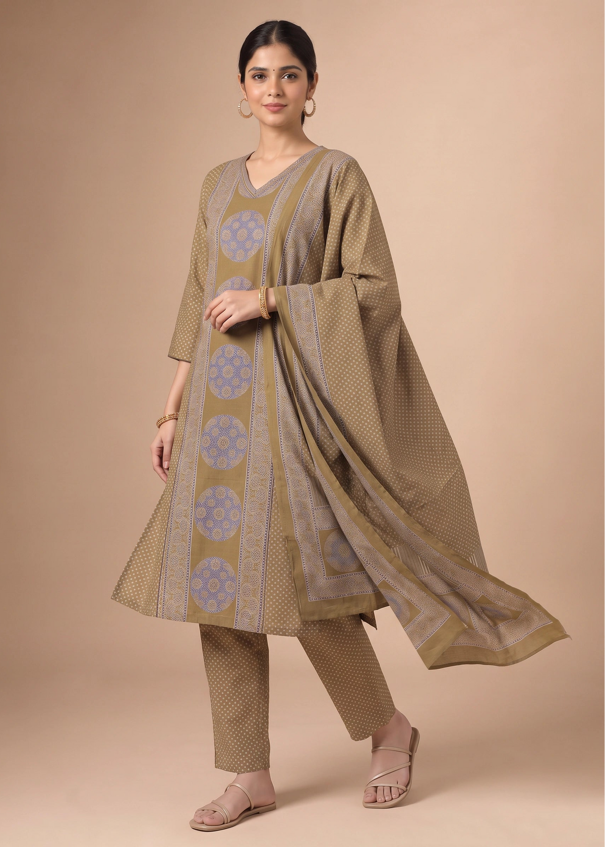 A woman models a traditional Indian salwar kameez suit set in a studio setting. The set features a taupe-beige kurta and trousers with light blue geometric patterns, paired with a matching sheer dupatta.