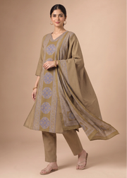 A woman models a traditional Indian salwar kameez suit set in a studio setting. The set features a taupe-beige kurta and trousers with light blue geometric patterns, paired with a matching sheer dupatta.
