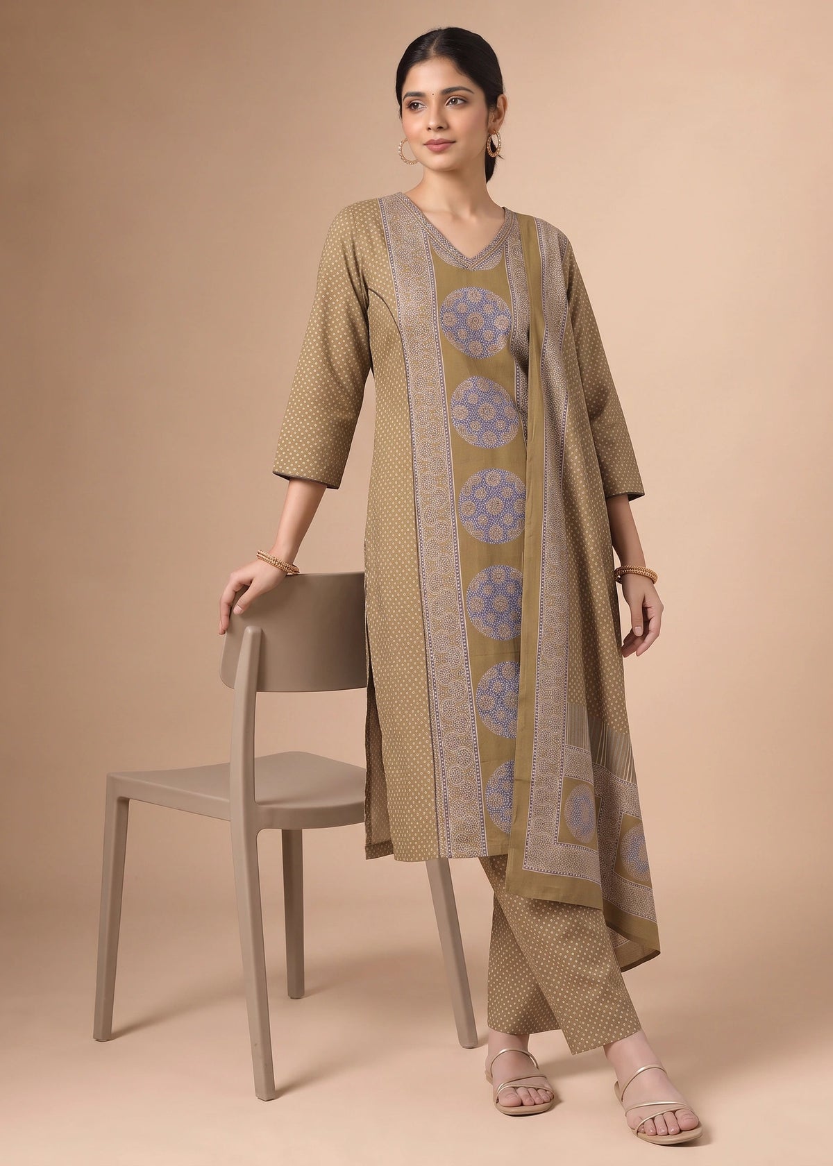 A woman models a beige and light blue printed kurta set with matching pants, standing next to a matching beige chair against a solid beige background.