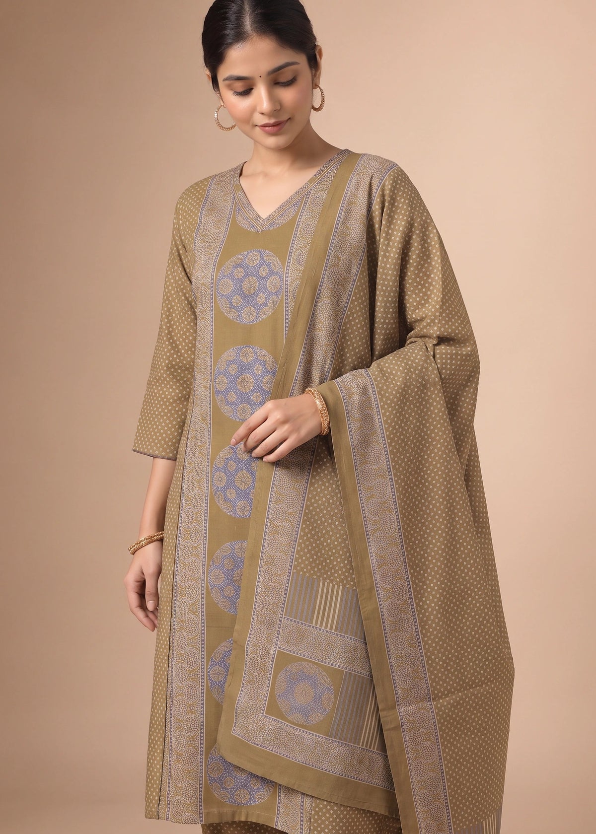 A woman models a traditional South Asian salwar kameez suit set in a warm khaki-beige color, featuring light blue and beige geometric patterns on the kurta and dupatta, set against a solid beige studio background.