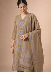 A woman models a traditional South Asian salwar kameez suit set in a warm khaki-beige color, featuring light blue and beige geometric patterns on the kurta and dupatta, set against a solid beige studio background.