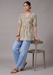 A woman models a beige and green patterned short-sleeve tunic top paired with light blue jeans and brown sandals, sitting on a minimalist taupe chair against a solid gray studio background.