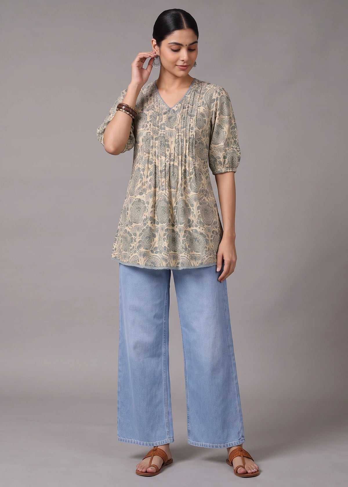A woman modeling a short-sleeve V-neck tunic top with a beige and green floral pattern, paired with wide-leg light blue jeans and brown sandals, against a solid gray studio background.