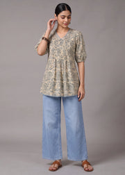 A woman modeling a short-sleeve V-neck tunic top with a beige and green floral pattern, paired with wide-leg light blue jeans and brown sandals, against a solid gray studio background.