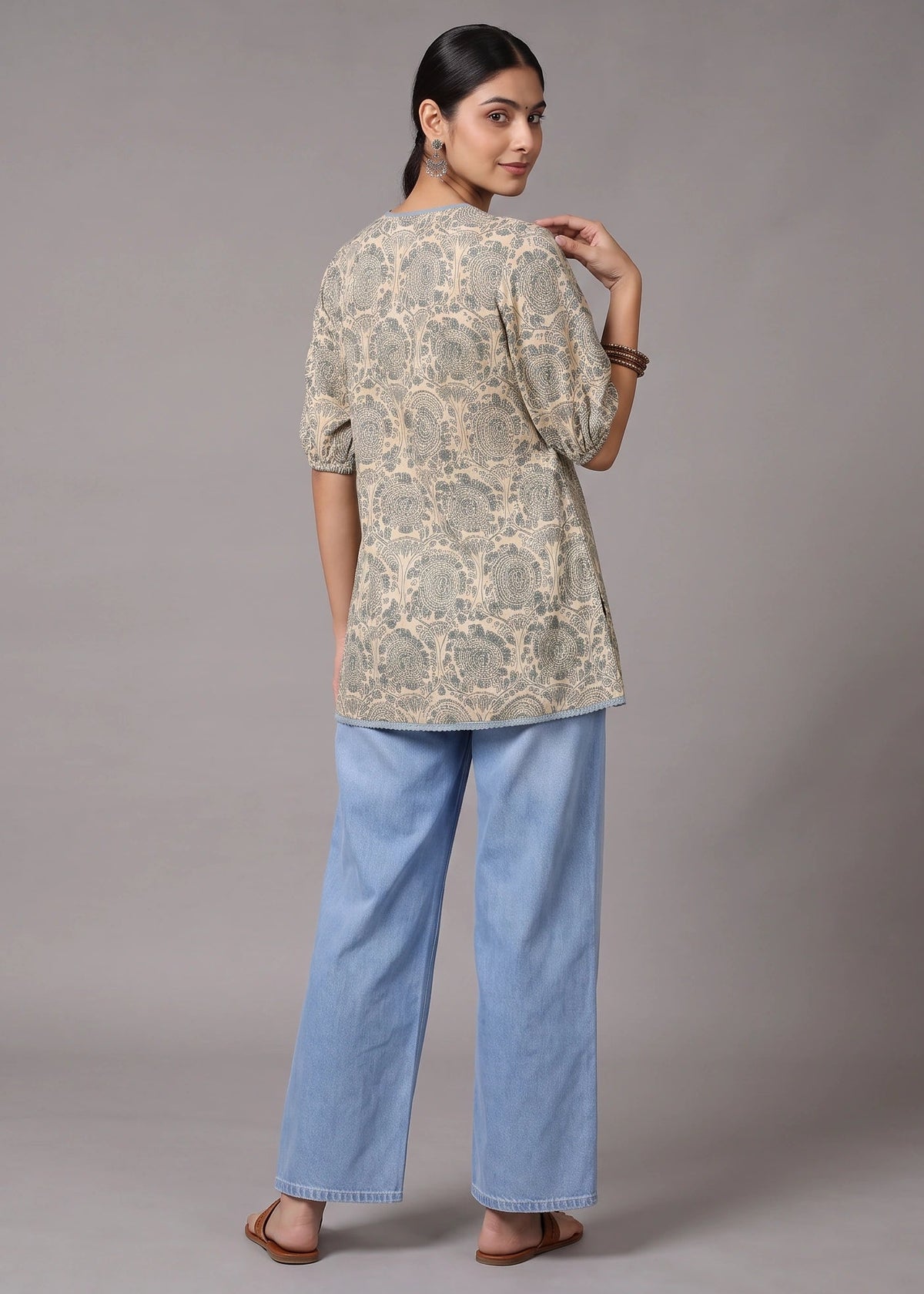 A woman is shown from the back wearing a beige and gray patterned tunic top with light blue wide-leg jeans, standing against a neutral gray studio background.