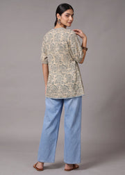 A woman is shown from the back wearing a beige and gray patterned tunic top with light blue wide-leg jeans, standing against a neutral gray studio background.