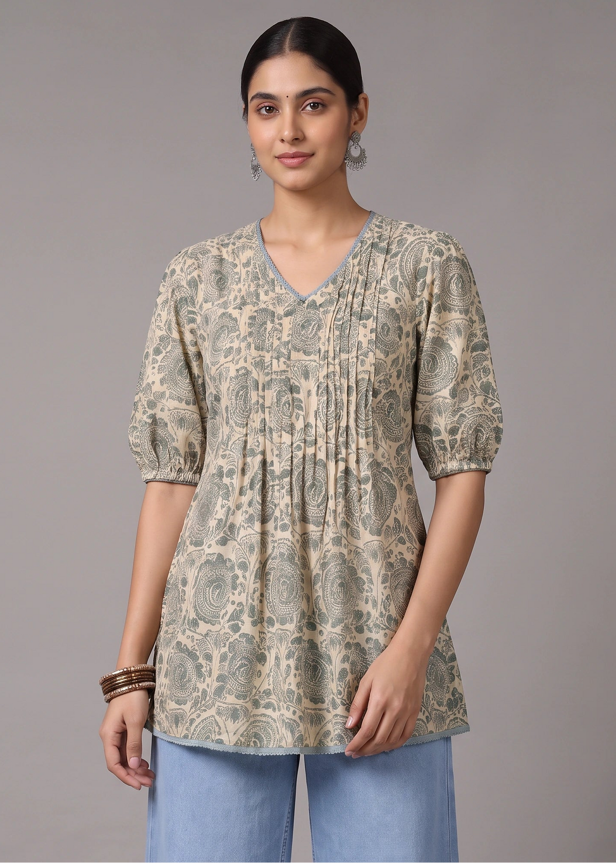 A woman modeling a short-sleeve V-neck tunic top with an intricate beige and gray floral block print, paired with light blue jeans, against a solid gray studio background.
