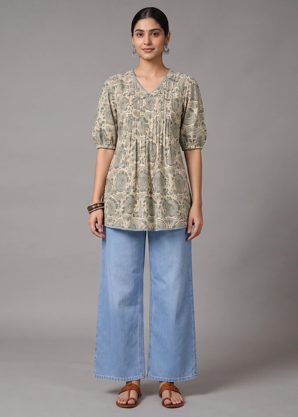 A woman stands against a plain gray studio background wearing a short-sleeve V-neck tunic top with an abstract beige and olive print, paired with wide-leg light blue jeans and brown sandals.