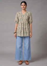 A woman stands against a plain gray studio background wearing a short-sleeve V-neck tunic top with an abstract beige and olive print, paired with wide-leg light blue jeans and brown sandals.