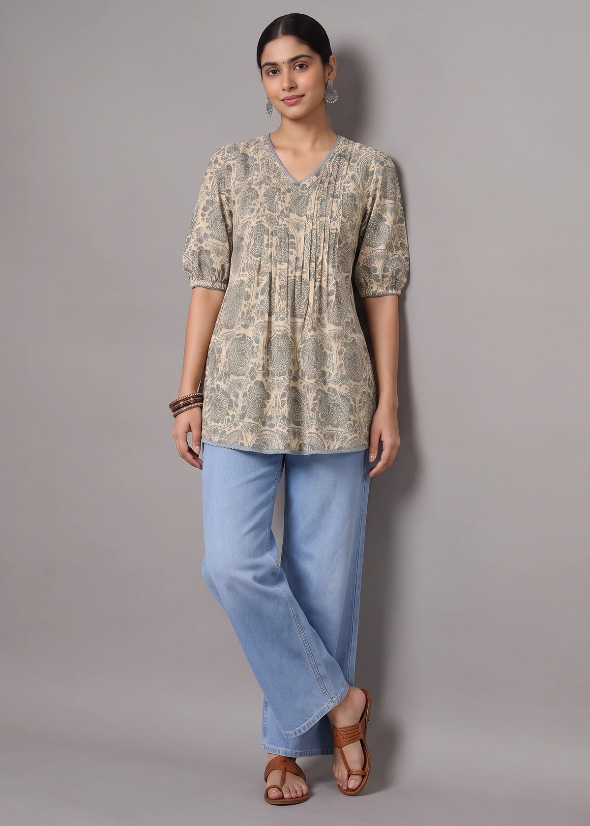 A woman modeling a beige and green floral print tunic top with puff sleeves, paired with light blue wide-leg jeans and brown sandals, standing against a solid gray studio background.