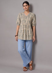 A woman modeling a beige and green floral print tunic top with puff sleeves, paired with light blue wide-leg jeans and brown sandals, standing against a solid gray studio background.
